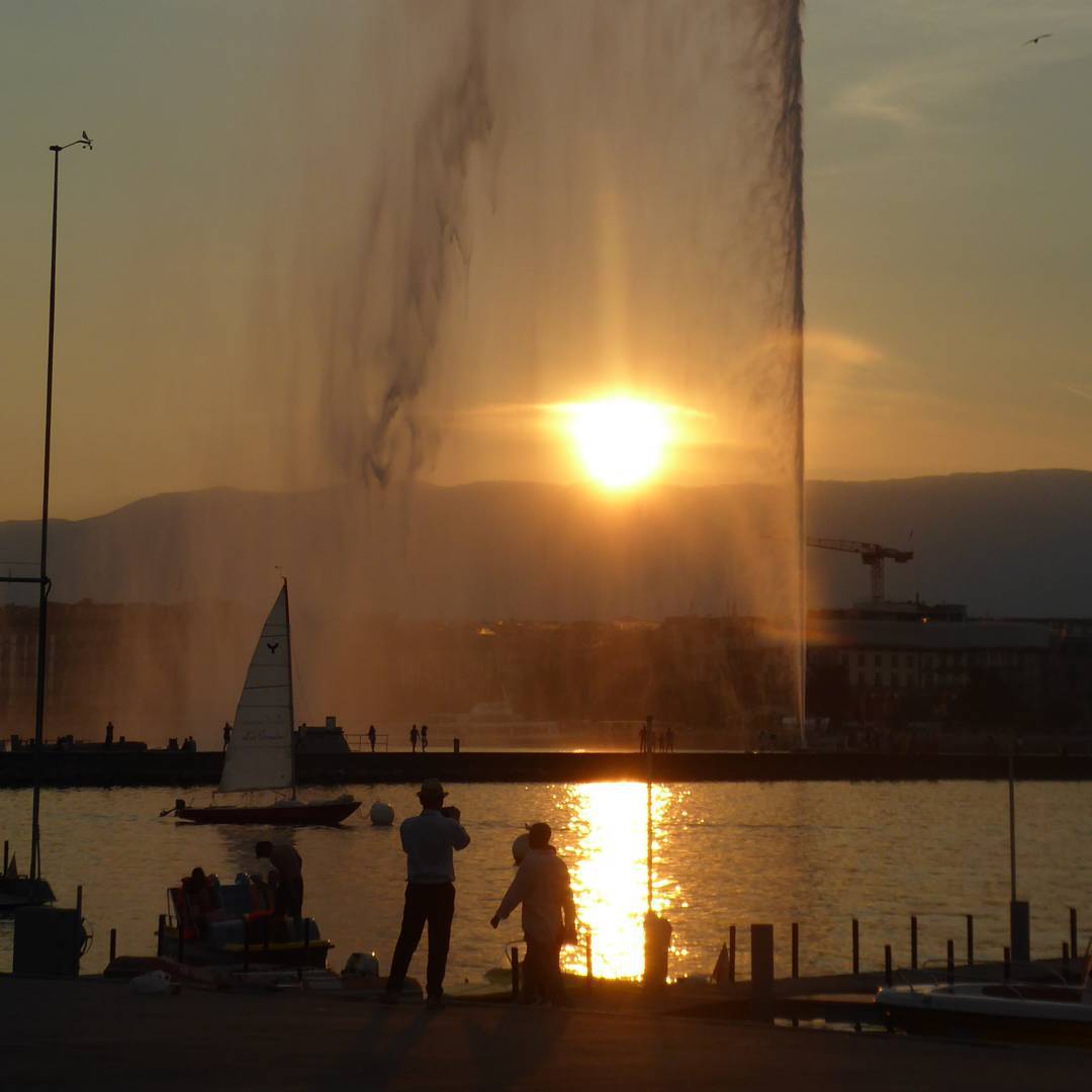 As the sun sets and blacklights the jet d'eau a groups of people get take away pizza and eat them by the lake side in geneva.
